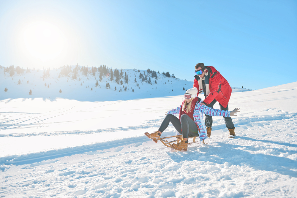 A couple enjoying a sled ride on a wooden sledge with the woman lifting her hands up and the man pushing the sledge