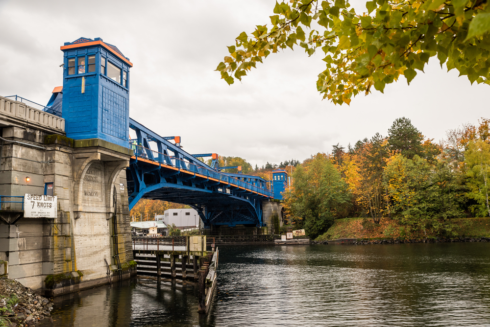 Colorful blue and orange drawbridge over a Seattle waterway with fall foliage trees in the background on a gray sky day.