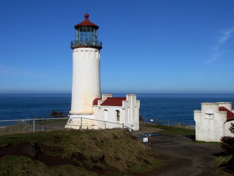 A white lighthouse with red detaiing next to a calm ocean on a sunny blue sky day.