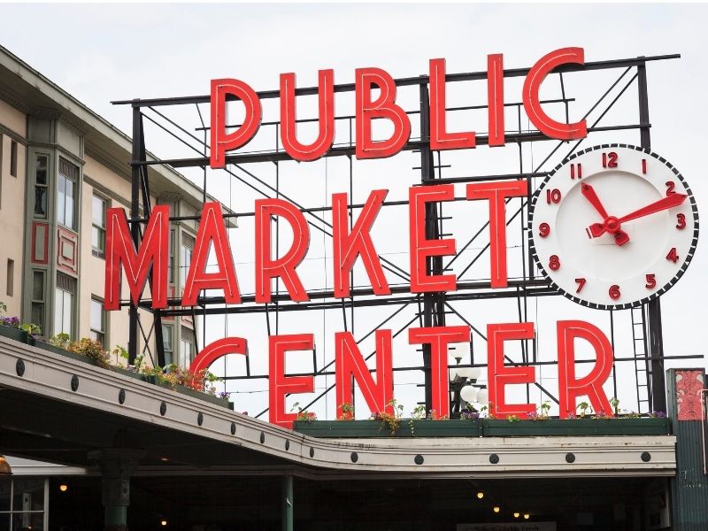 Vintage lettering which reads "Public Market center" in all-caps red font with a clock, also white and red, at the entrance to Pike Place Market, one of the top things to do in Seattle for foodies.