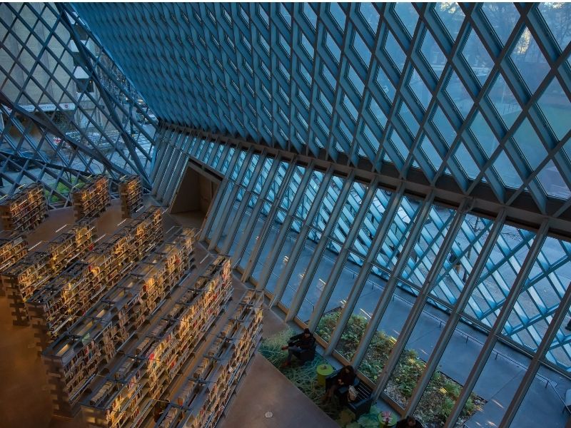 Photo from a higher floor of the Seattle Public Library showing the glass and steel diamond-pattern construction as well as shelves upon shelves of books on display.