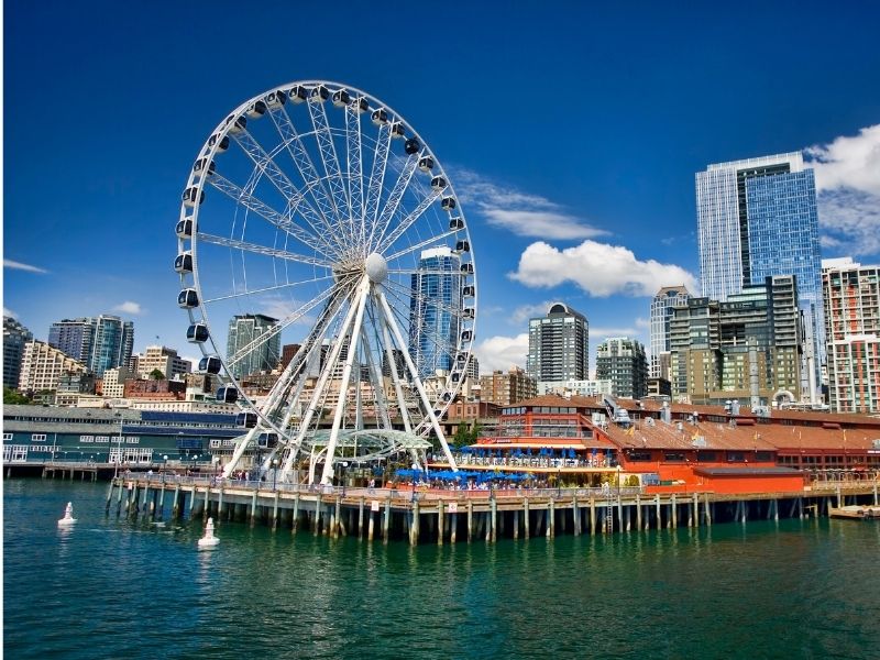 The Seattle Ferris Wheel on a sunny day on a pier with brilliant turquoise water, skyline in the background, and a partly cloudy sky.