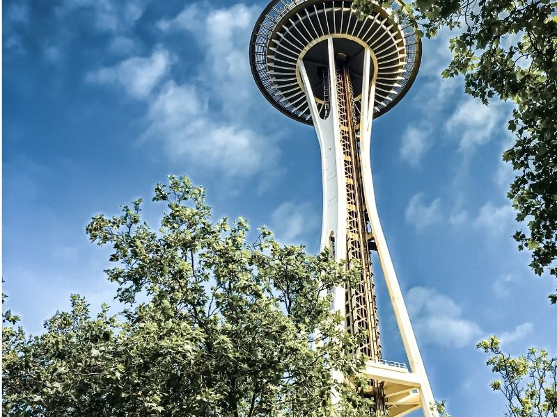 View from below of the Space Needle with trees framing the photo and a partly cloudy blue sky.