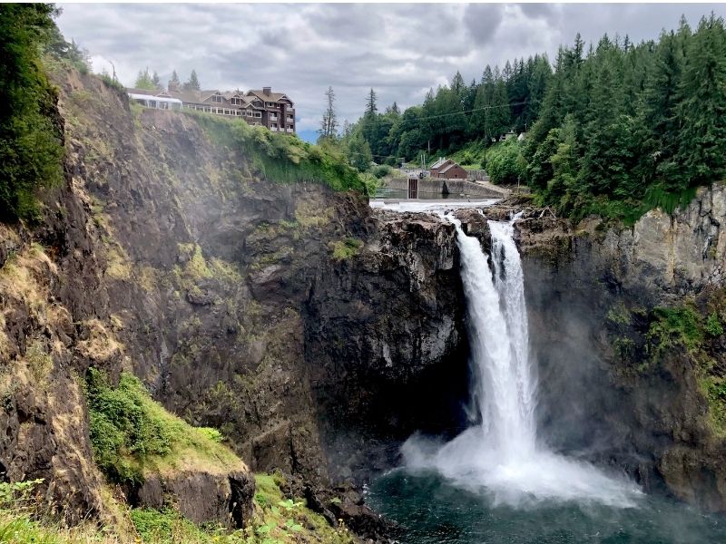 View of Snoqualmie Falls waterfall on a cloudy day with water thundering over into a turquoise pool below and pine trees framing the waterfall.