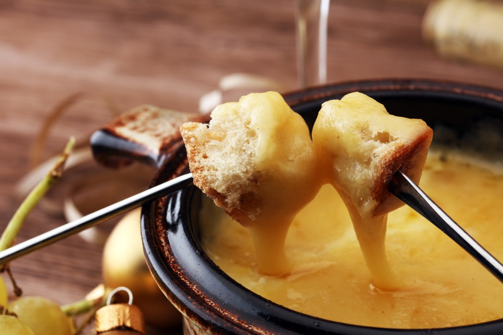 Two cubes of bread being dunked in hot melted cheese in a fondue pot using fondue forks.