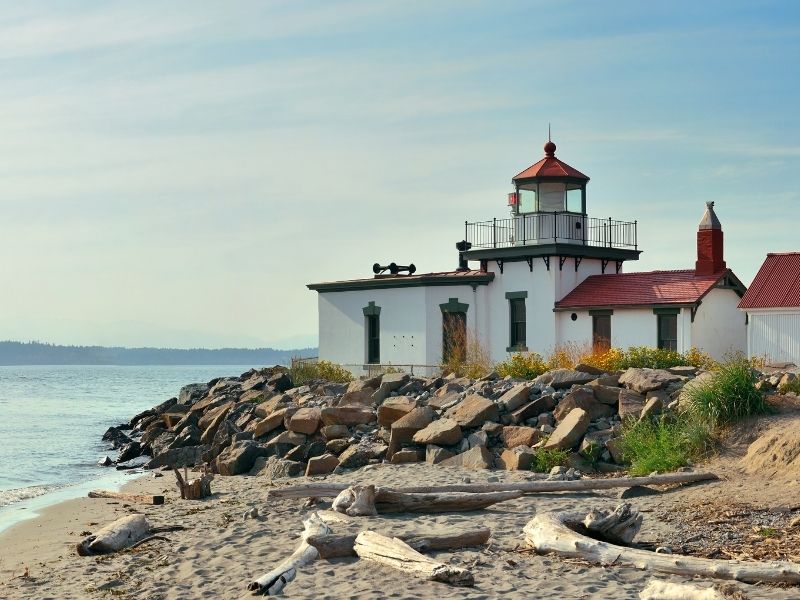 A sandy shore with some driftwood and large rocks with a lighthouse with white walls, and dark green and red detailing on it.