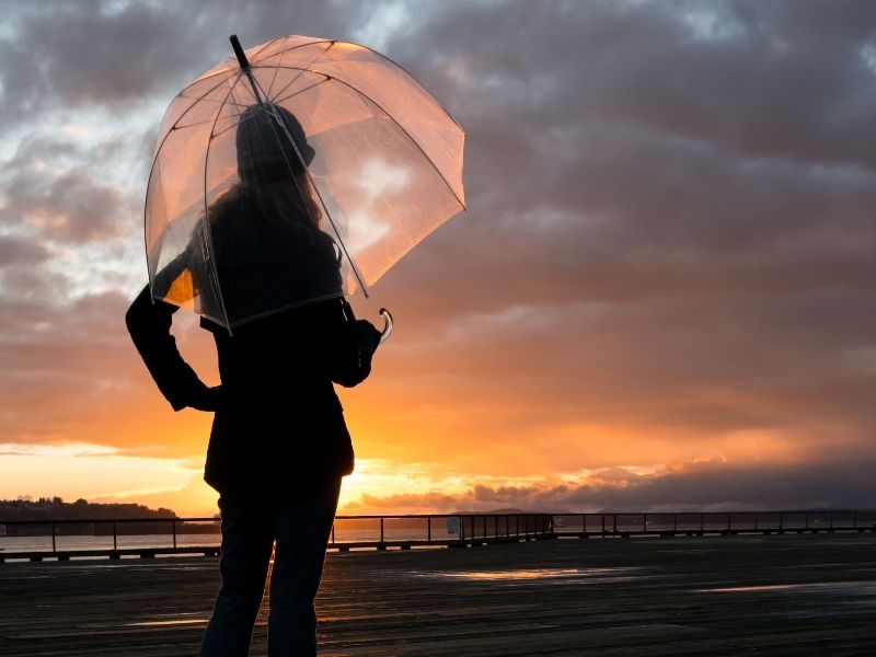 A woman carrying a clear umbrella on a rainy, overcast day in Seattle while the sun sets in the distance: an umbrella is a must-have Seattle packing list item!