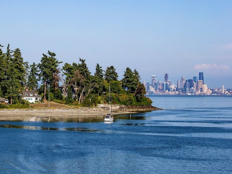 View from Bainbridge Island ferry of the Puget Sound waters, a house on an island with a sailboat in front, and in the distance, the skyline of Seattle.