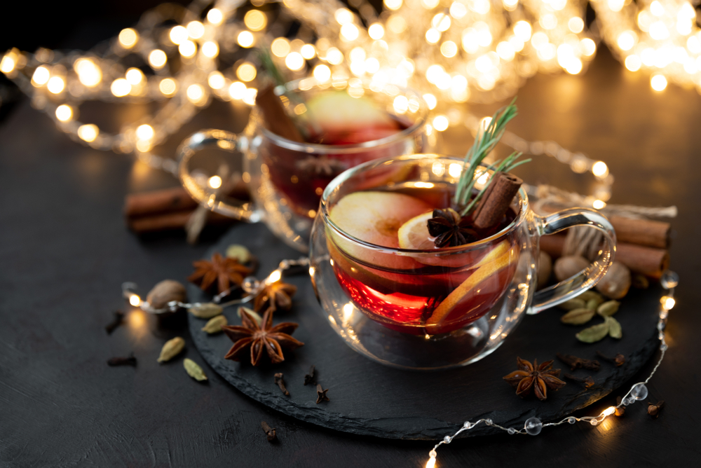 Two cups of mulled wine with spices used in making mulled wine around them with Christmas lights out of focus in background.