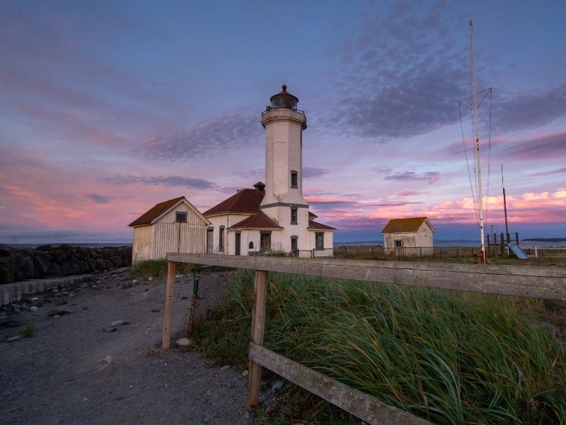 Sunset at Point Wilson lighthouse with a pink and purple sky lighting up the white and red-roofed lighthouse on the coastline.