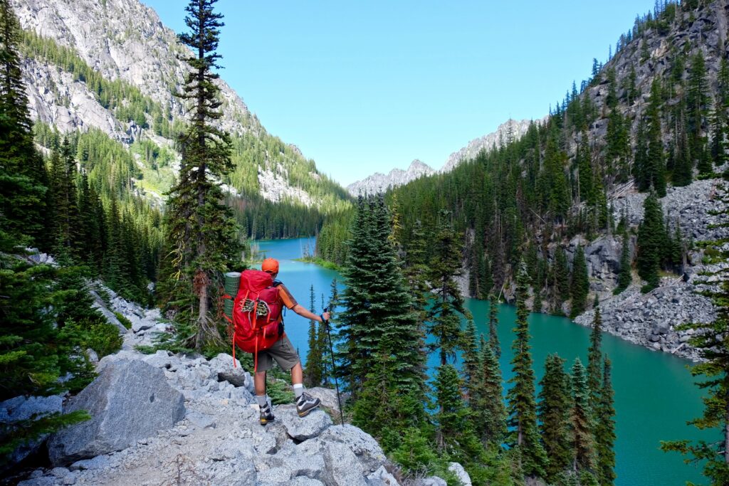 man carrying a large red backpack standing in front of a bright blue lake in washington state when hiking near seattle