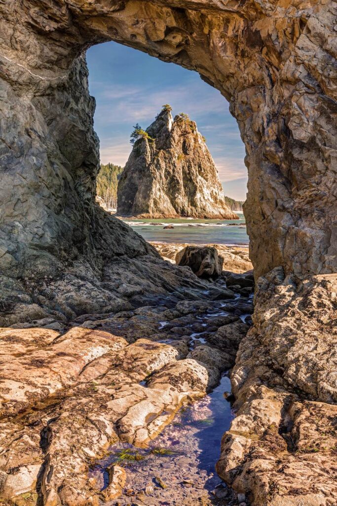 A natural rock arch on the beach that perfectly frames a tall sea stack rock formation behind it.