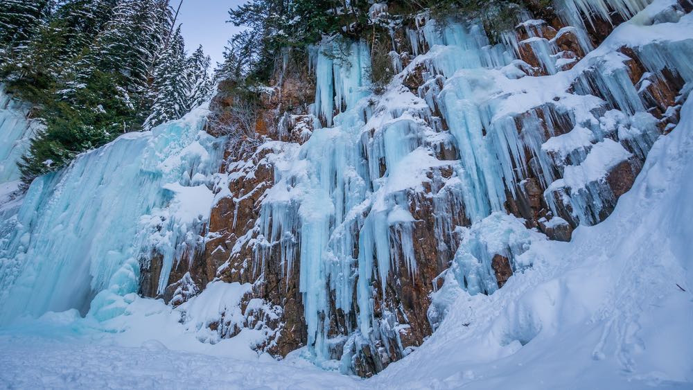 A frozen waterfall with giant pale blue icicles dropping down a cliff face, with snow at the base of the frozen waterfall. Franklin Falls is a beloved hike in the winer in Washington.