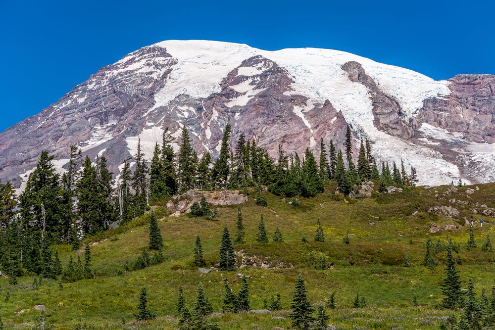 Grass-covered fields at the base of a large mountain cap with some teeny hikers in the right side corner for perspective.