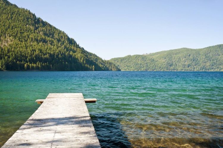 A small wooden pier leading out to brilliant blueish-turquoise lake water, surrounded by pine-tree covered mountains.