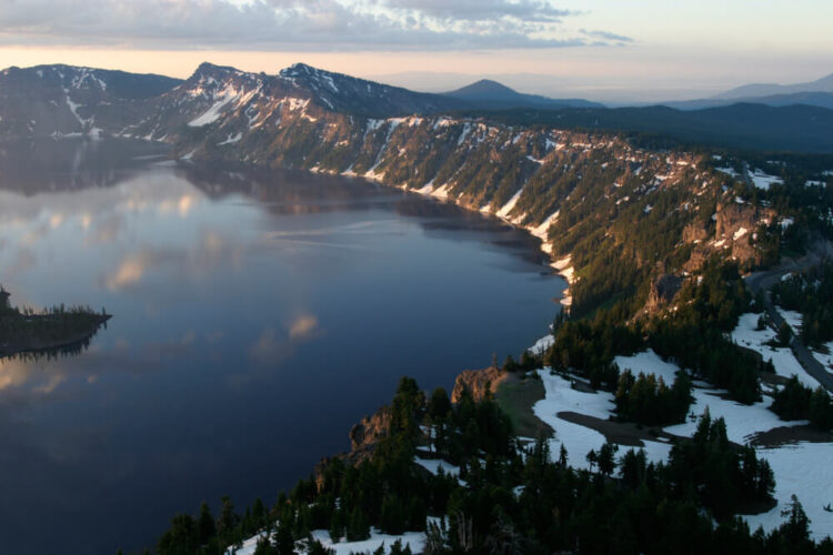 View of the lake from up on the caldera rim, with snow and trees and a view of a bit of the island in the middle of Crater Lake, with some light snow on the ground in winter. Sunrise or sunset.