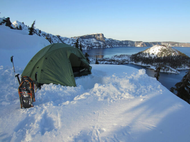 A tent perched in the snow, next to a pair of of snowshoes, overlooking Wizard Island covered in snow in the middle of Crater Lake in winter.