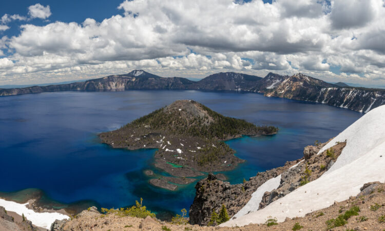 View of the island in the middle of Crater Lake, Wizard Island, surrounded by blue and teal water, seen from an overlook covered in light snow.