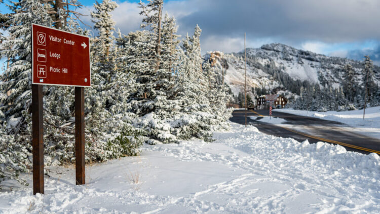 A sign reading "Visitor Center, Lodge, Picnic Hill" surrounded by snow, a plowed road leading to buildings off in the distance.