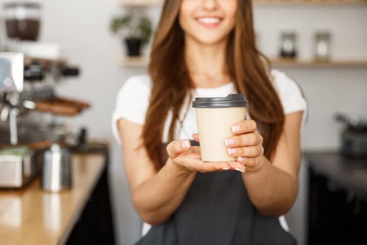 young woman working at a bend or coffee shop holding out a to-go cup of coffee