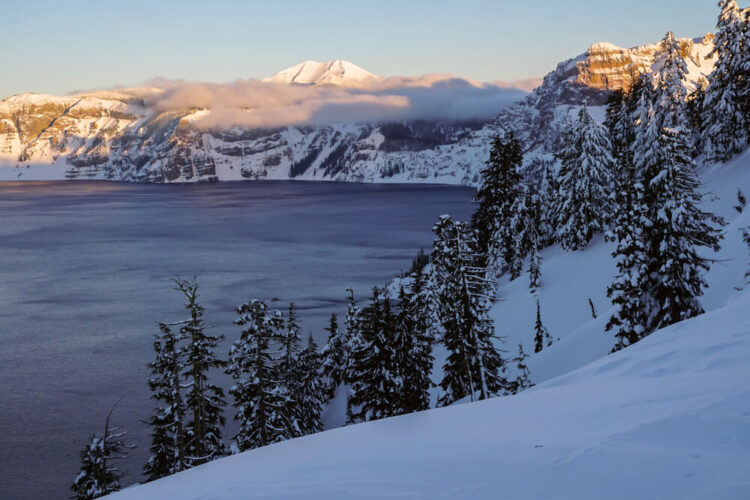 View of Crater Lake at sunrise, with snow-covered rim, some low-lying clouds on part of the caldera, and alpenglow (pink tinged peaks) from the morning sun on a few select ridges.