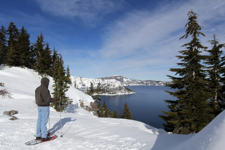 A man in a hoodie and jeans wearing snowshoes looking out onto a snow-covered Crater Lake winter landscape, including Wizard Island covered in snow, on a sunny day.