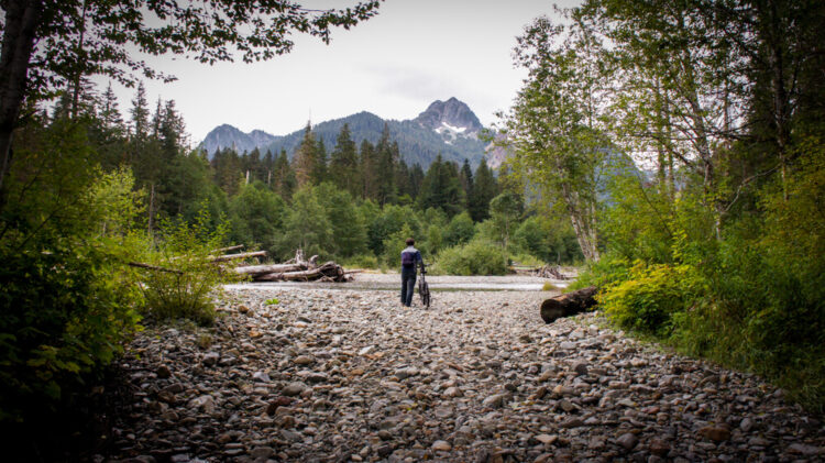 Man with a mountain bike walking towards a mountain in the distance, on a rocky path, preparing to cross a stream.