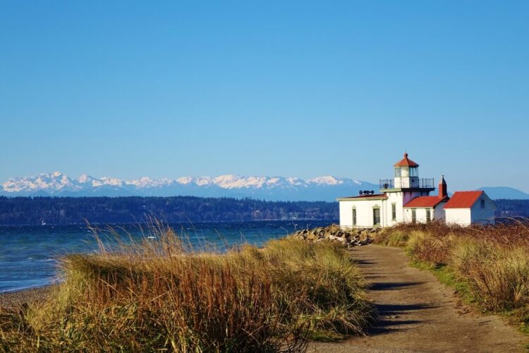 A grassy path leading to a lighthouse in Seattle on the water with mountains in the background.