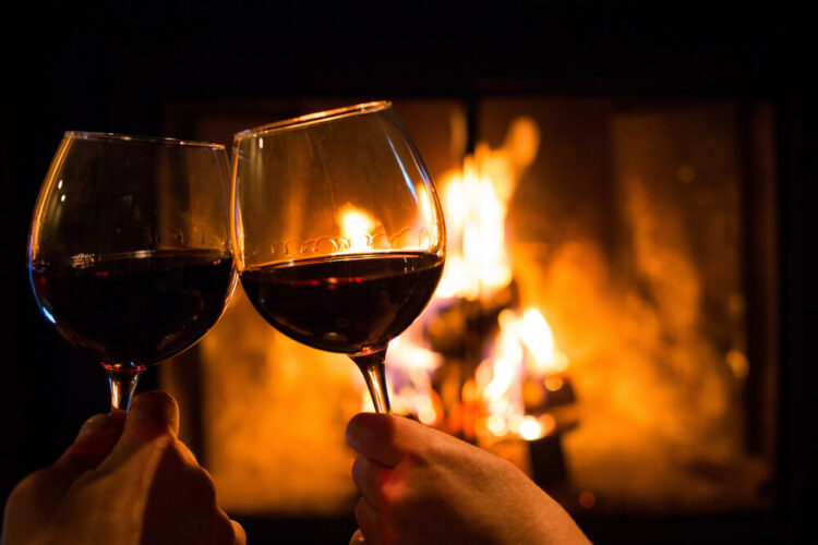 Two glasses of red wine clinking in 'cheers' in front of a roaring fireplace.