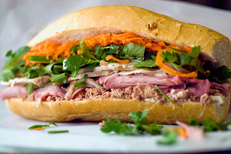 A close up photo of the most traditional style of Banh Mi, with pate, roast pork, and cold cuts with cilantro, carrot, and other fixings on a rice flour baguette.