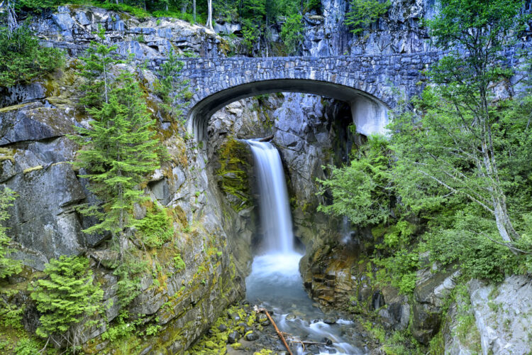 A beautiful single drop waterfall in Mt Rainier National Park, with a stone bridge in front of the waterfall, and trees around it.