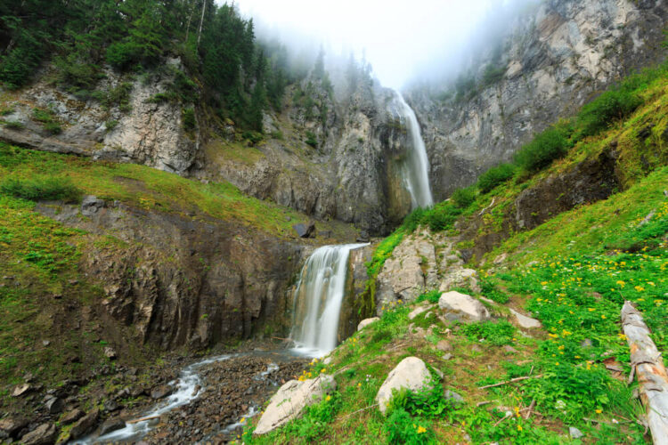 A two-tiered waterfall on a foggy day in mt Rainier National Park, surrounded by grass, logs, and stones.