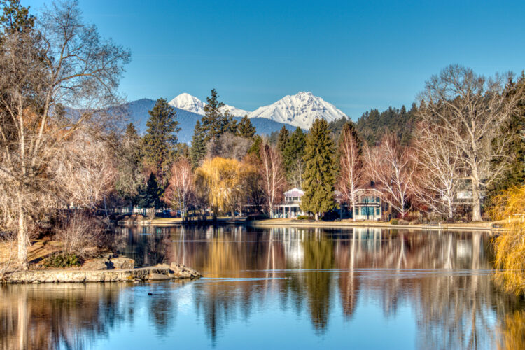 Glassy Mirror Pond in Drake Park surrounded by fall foliage and bare trees, with some houses or buildings around the pond, a popular Bend hike (or walk!)