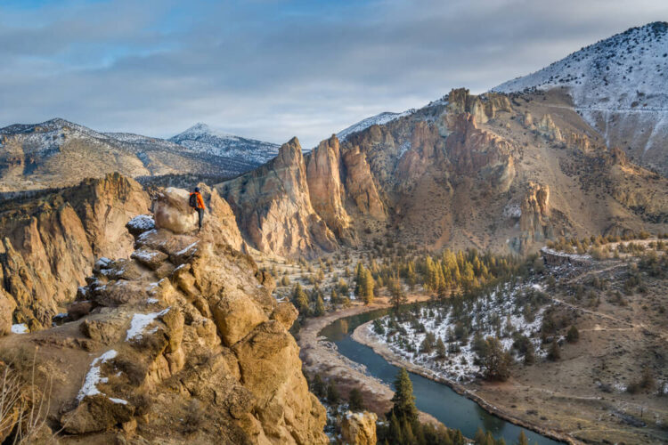 Hiker in a red shirt and black pants with a backpack on, looking over the landscape in Smith Rock, Oregon over a river with a light dusting of snow.