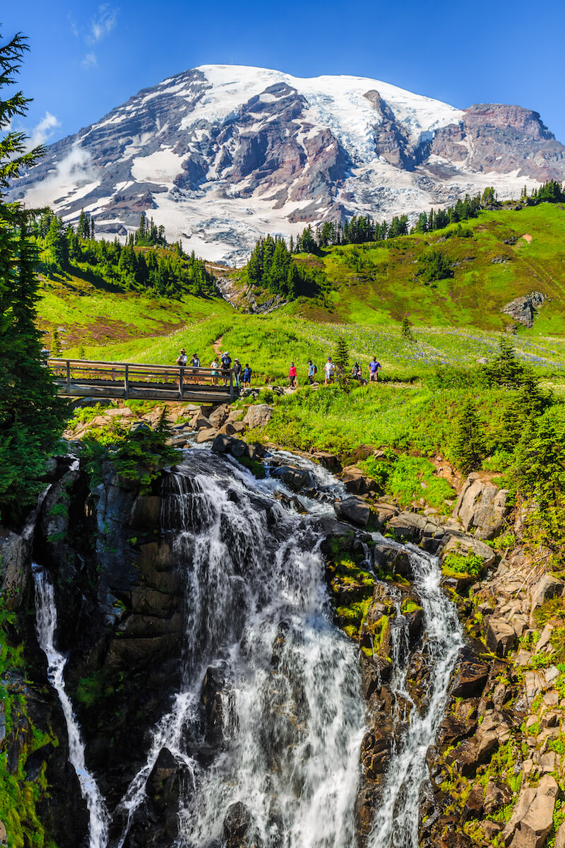 A group of hikers crossing a bridge, which spans over a narrow but long waterfall in Mt Rainier National Park, with a snow-capped mountain behind it.