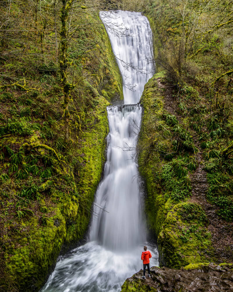 A man in a red jacket standing at an outlook point in front of the multi-tiered Bridal Veil Fall, one of the most famous waterfalls near Seattle