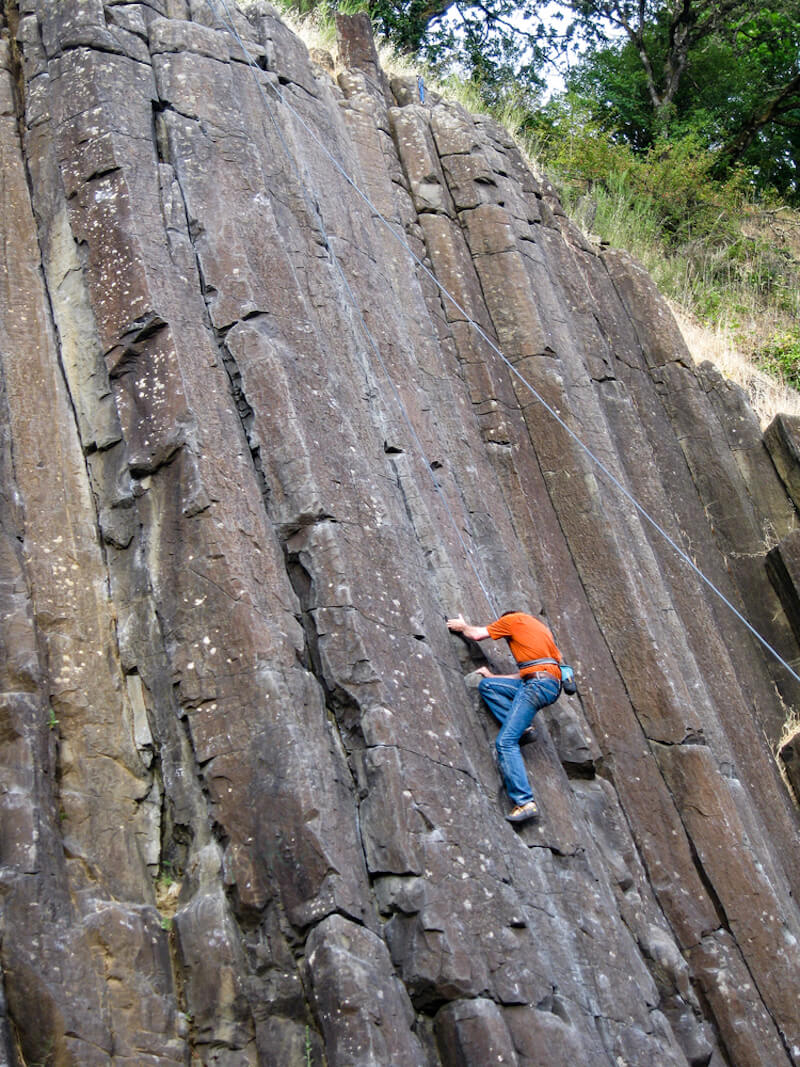 Man in orange shirt and jeans trying to climb up the basalt columns at Skinner Butte, a popular outdoorsy thing to do in Eugene Oregon