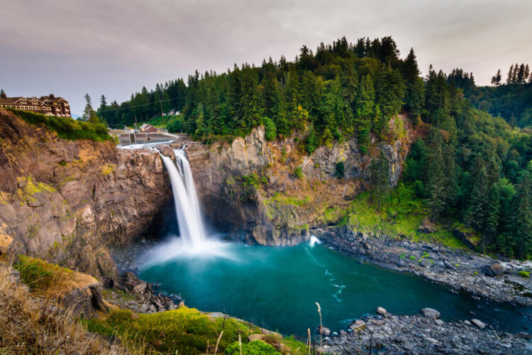 A waterfall tumbling off of a cliff edge into a turquoise pool of water, surrounded by trees, with a lodge overlooking this famous waterfall near Seattle on a cloudy day.