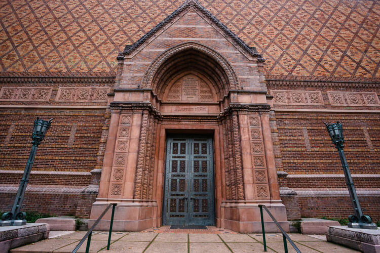 Brick red building with carving and two vintage looking lamposts with an elaborate door. Building is the University of Oregon Jordan Schnitzer Museum of Art building exterior on the Eugene Campus.