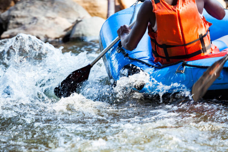 Black man in a orange vest in a blue raft going down white water rapids