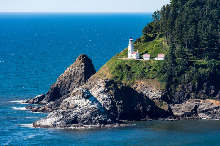 the white and red-roofed lighthouse of heceta head, next to rock formations and evergreen trees, against a backdrop of a sparkling blue pacific ocean