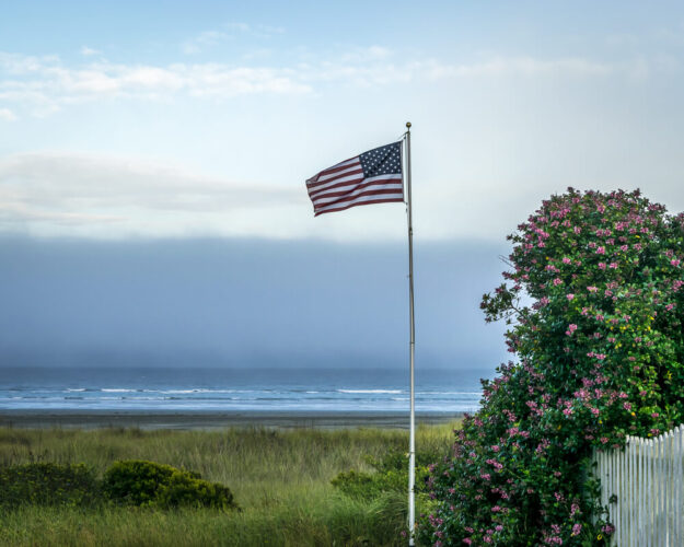 American flag waving in the wind surrounded by green grass and beach coastline with a white picket fence and pink flowers on the right of the photo.