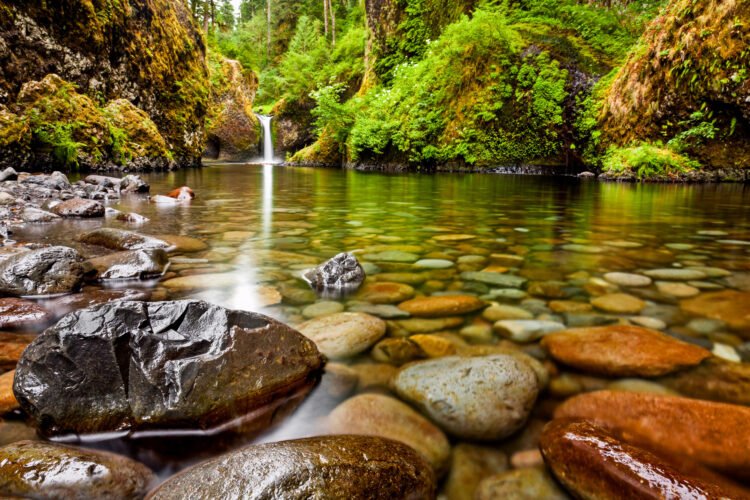 punch bowl waterfalls oregon as seen from a distance with a clear pool in the foreground, covered in stones