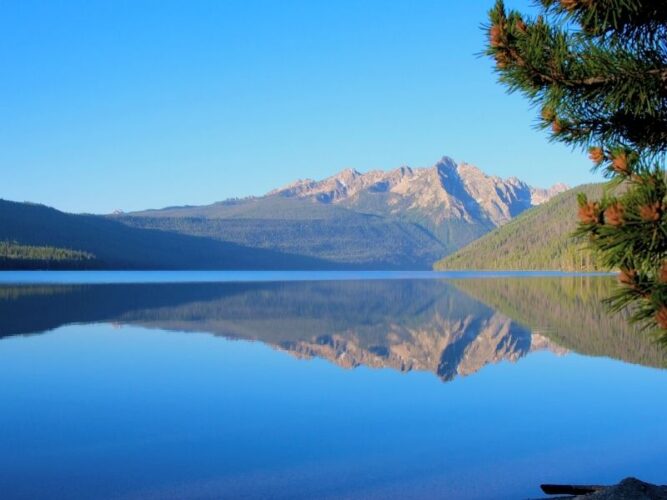 Clear, still blue waters reflecting the Sawtooth mountains with a bit of pine brush in the foreground, looking out onto a peaceful lake on an Idaho weekend getaway at Redfish Lake